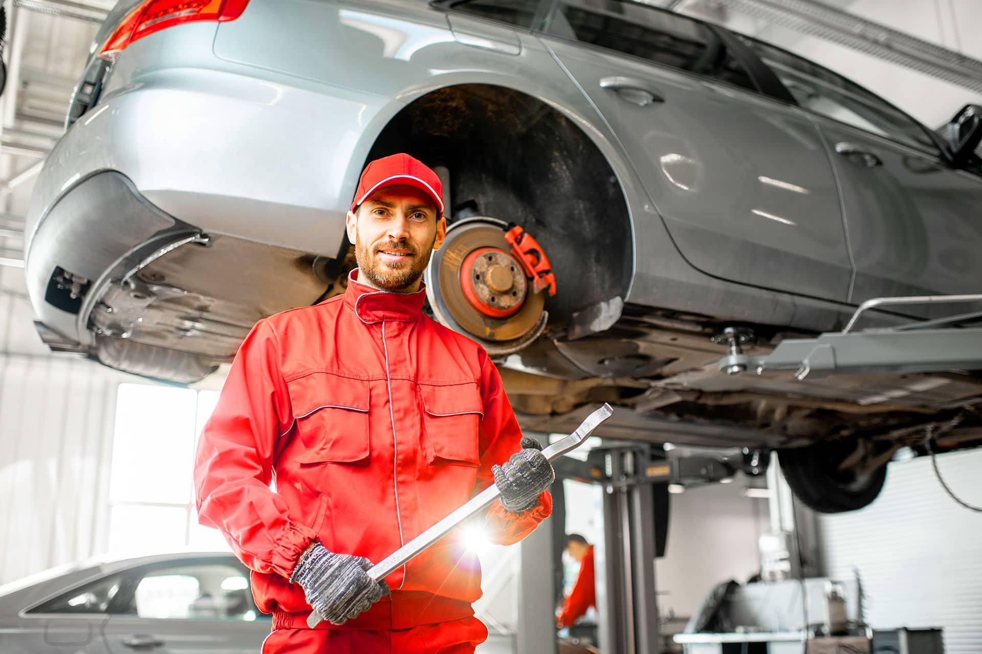 A mechanic in red coveralls holds a wrench while standing next to a car lifted on a hydraulic lift, showing the vehicle's wheel area. The workshop is well-lit, emphasizing the auto repair environment.