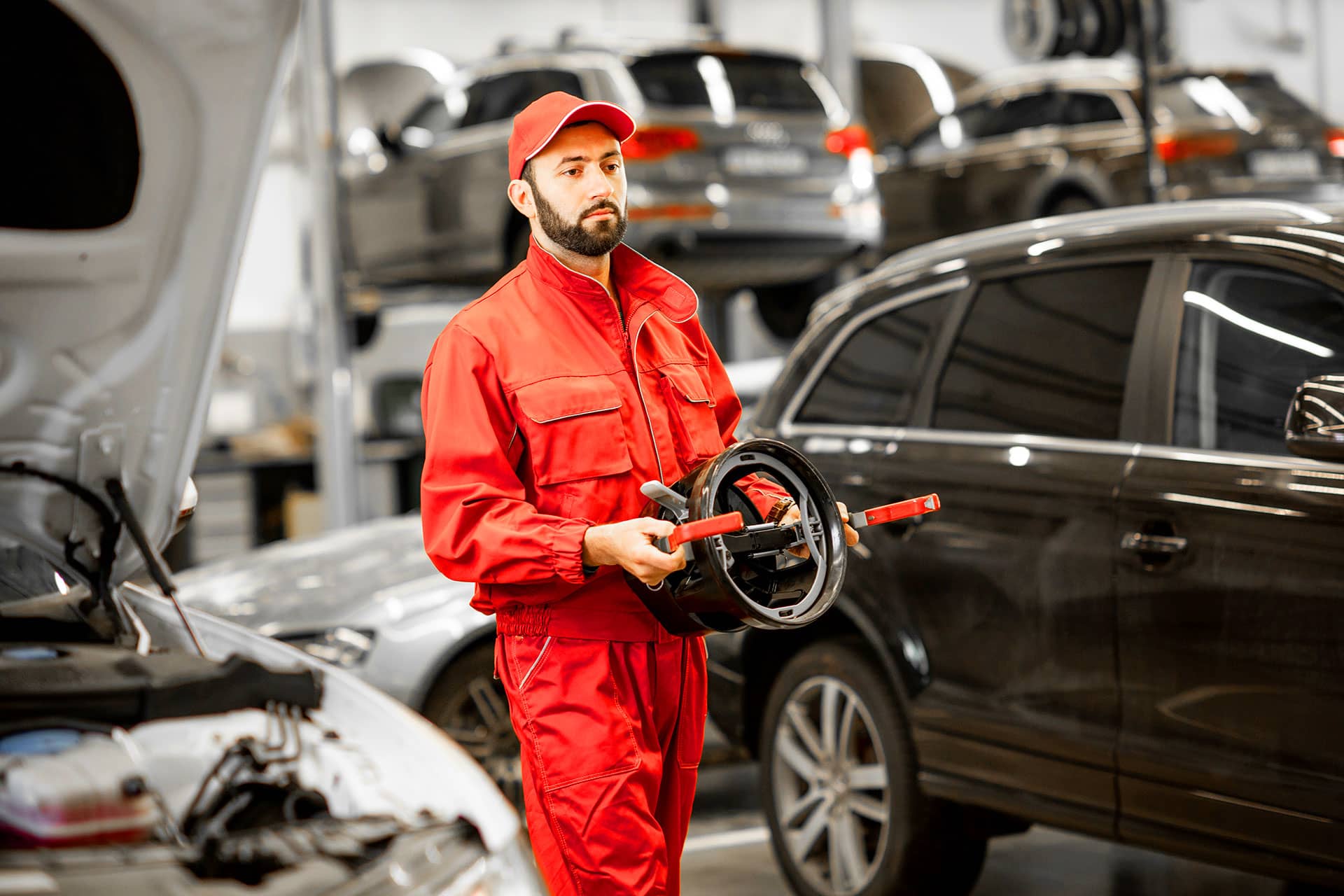 A mechanic in a red jumpsuit stands in an auto repair shop, holding a tool while surrounded by vehicles. The shop features open hoods and various cars in the background, indicating ongoing repairs.