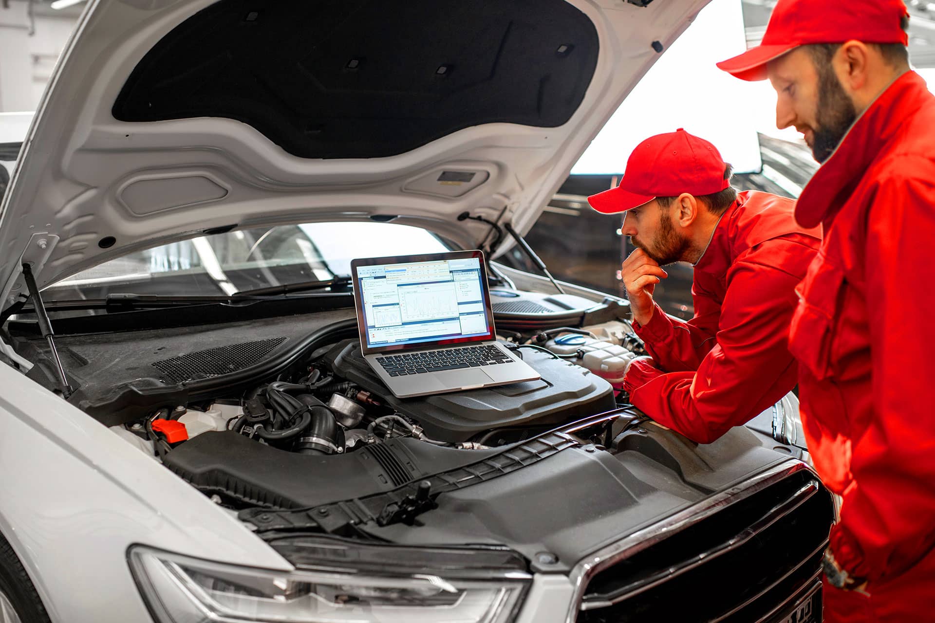 Two mechanics in red uniforms inspect a car's engine while using a laptop to diagnose issues. The vehicle's hood is open, revealing its engine components.